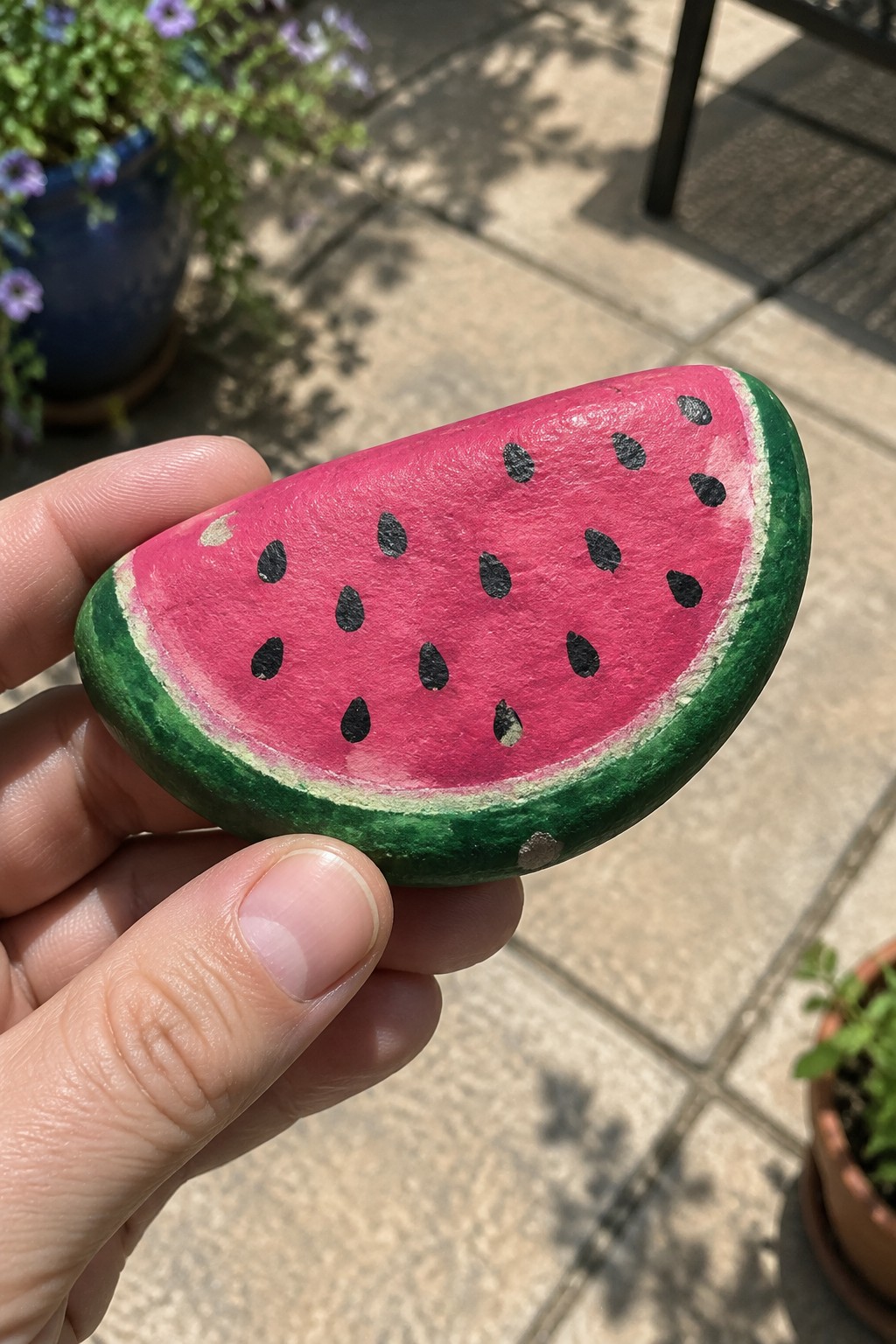 A hand holds a smooth oval rock painted to look like a watermelon slice with pink center, black seeds, and green rind.