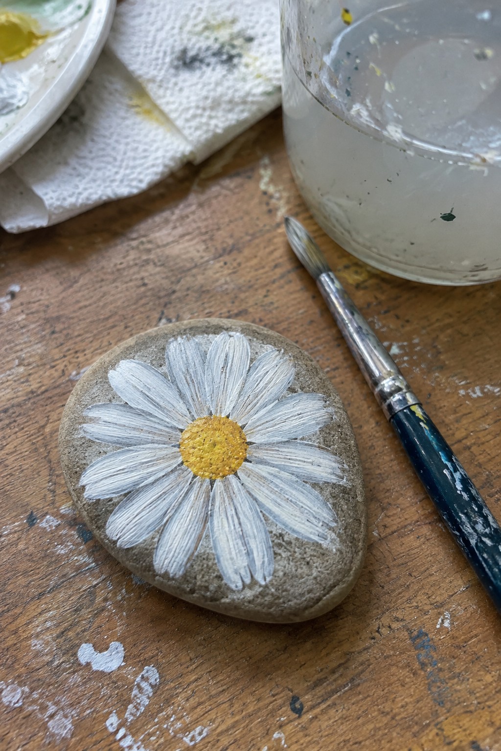 Painted rock with white daisy and yellow center beside paintbrush on wooden table