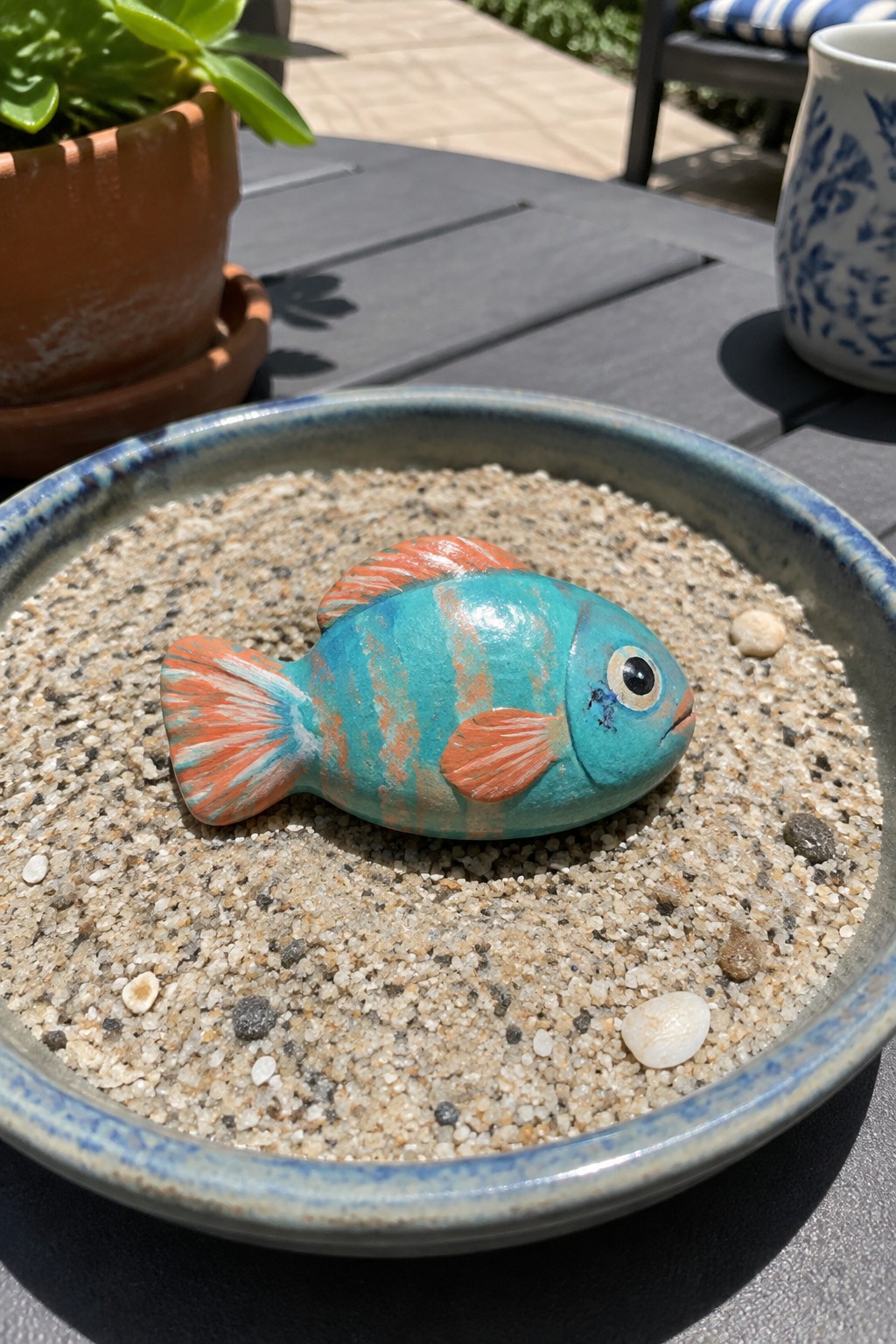 Painted turquoise fish with orange fins in a sand-filled bowl on table.