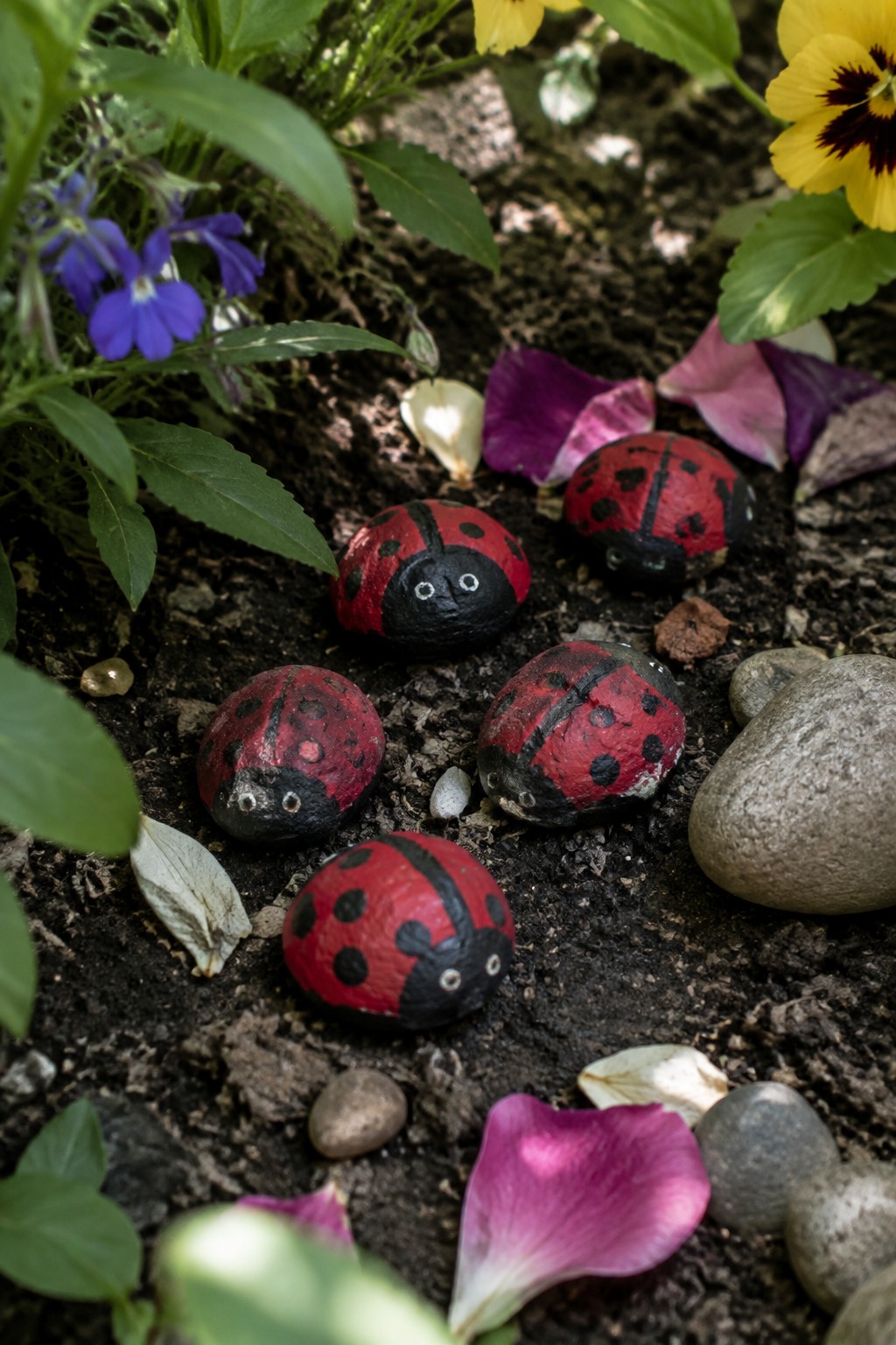 Painted ladybug rocks on soil amid flowers, petals, and leaves.