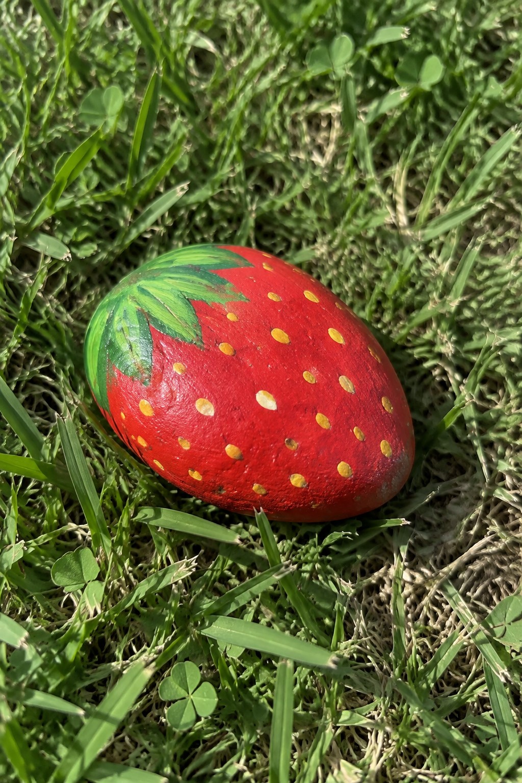 Red strawberry-painted rock with yellow dots and green leaves on grass