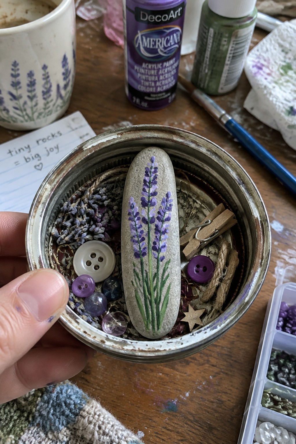 Hand holding metal tin with lavender-painted stone, buttons, dried flowers, and beads