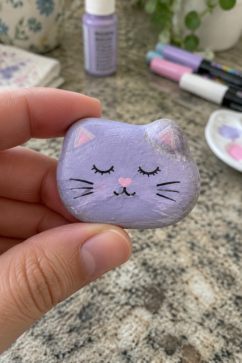 Hand holding purple painted cat rock with closed eyes, whiskers, and pink nose