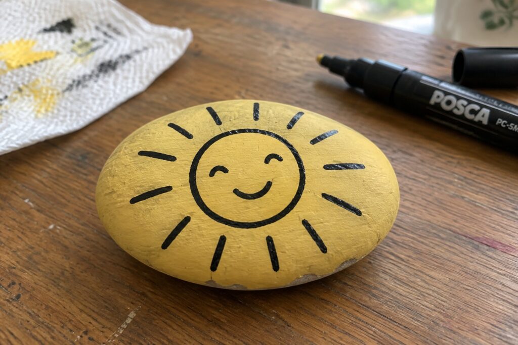 Yellow painted rock with smiling sun face beside black Posca marker on wood.