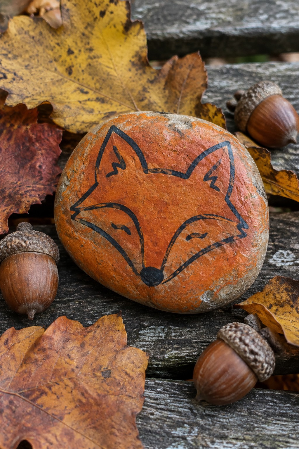 Orange rock painted with fox face amid autumn leaves and acorns on wood