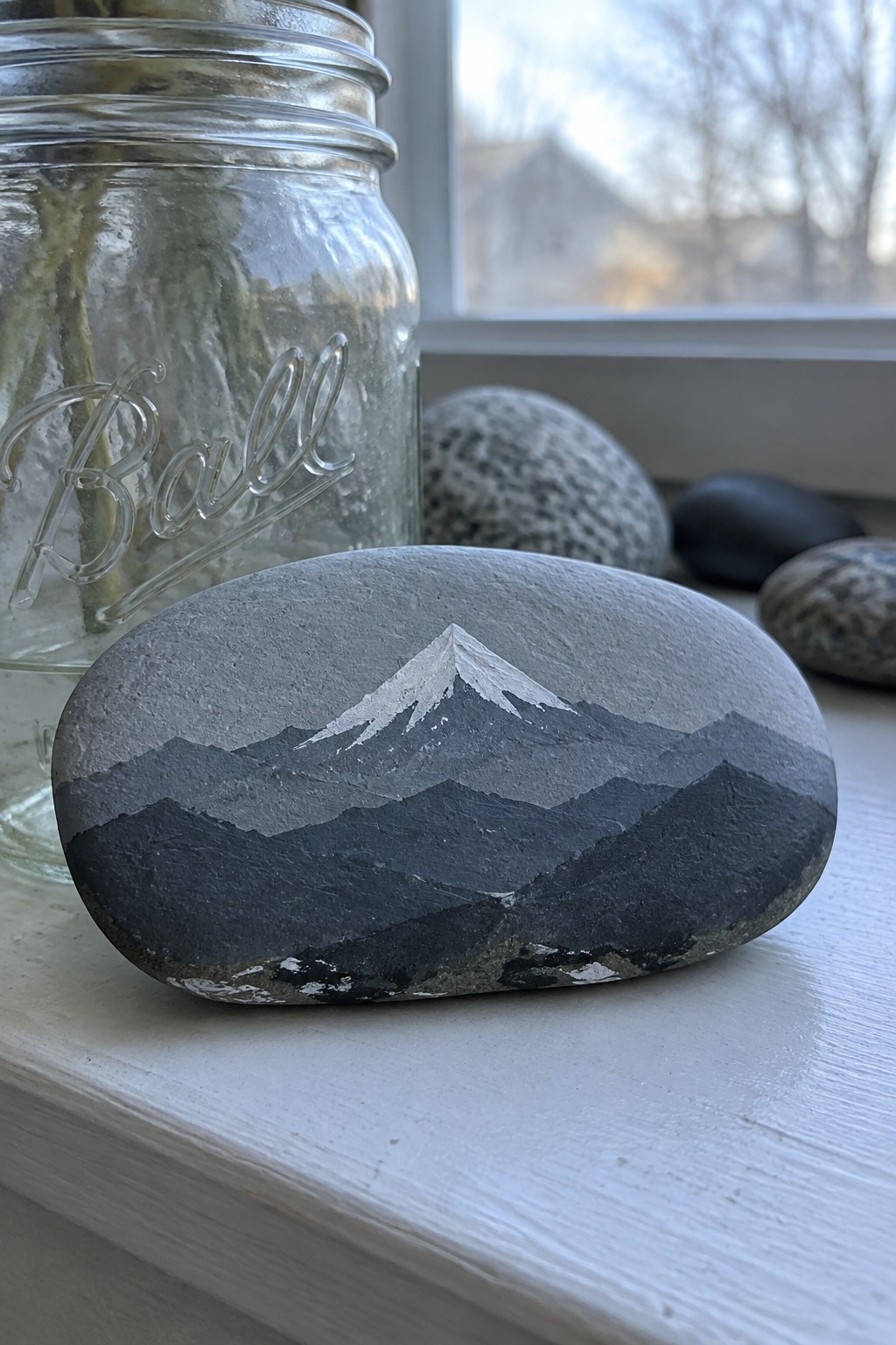 Oval rock painted with layered grayscale mountains beside Ball mason jar on sill
