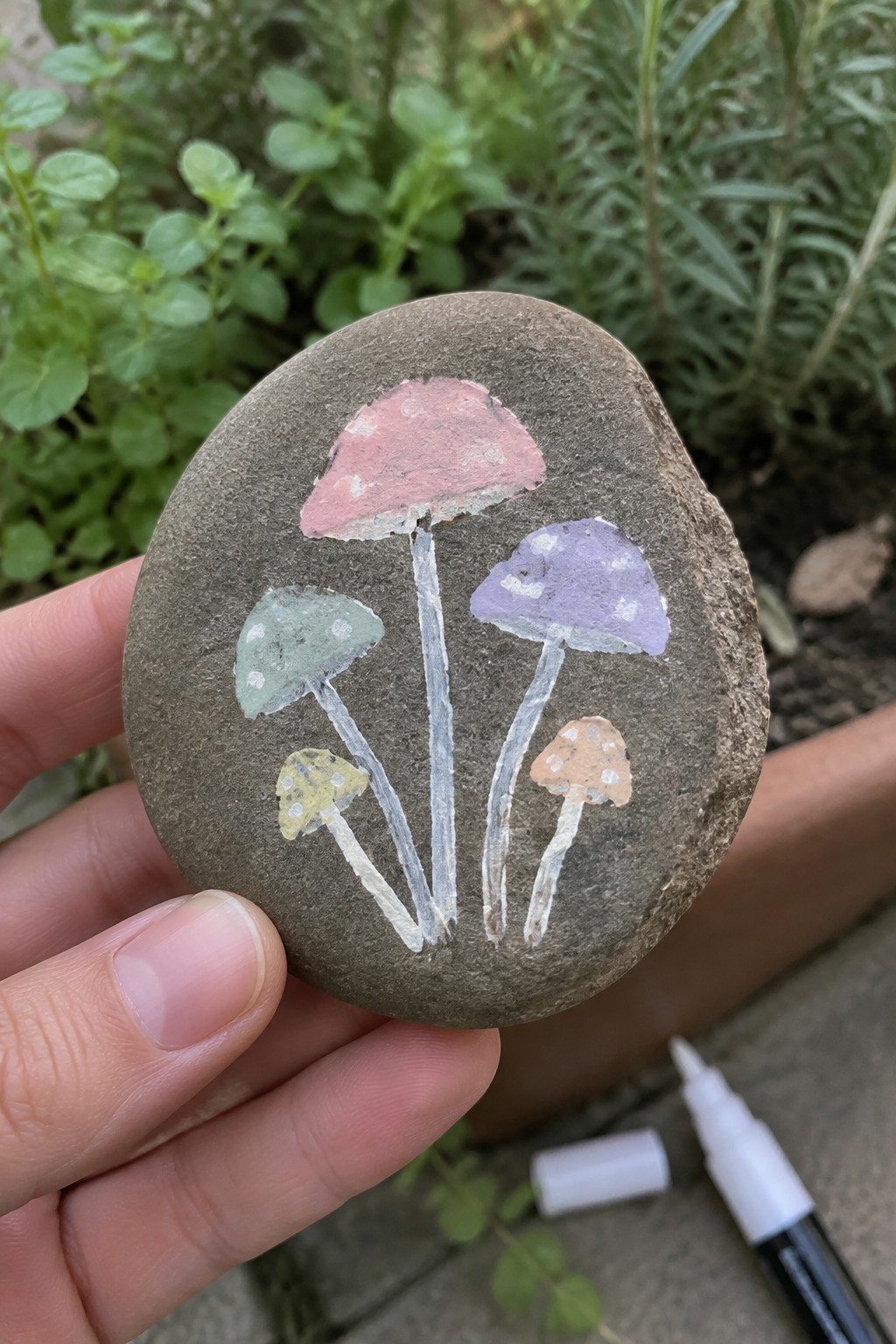 Hand holding gray rock painted with five colorful spotted mushrooms.