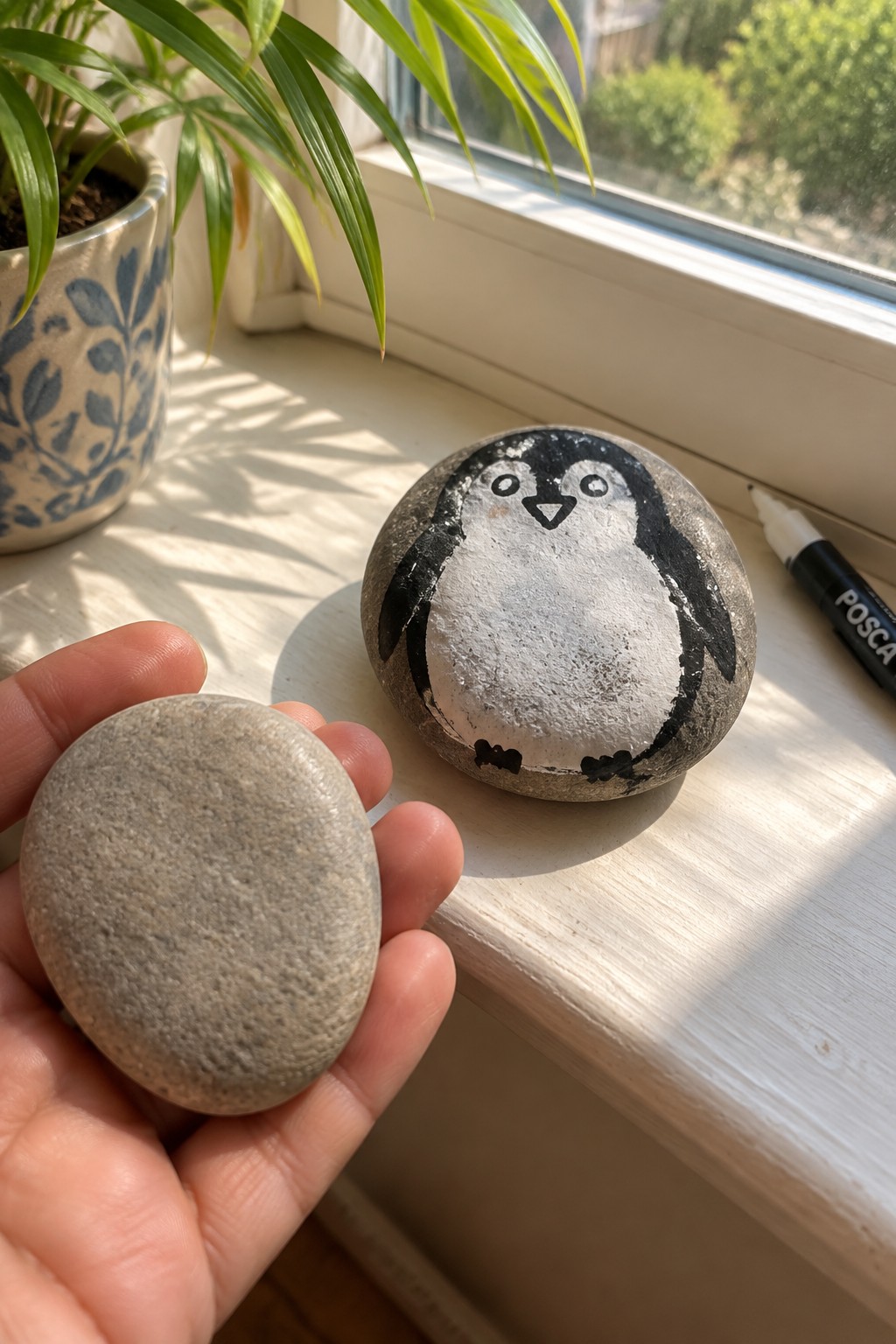 Hand holding plain gray stone near penguin-painted rock on sunlit windowsill with plant