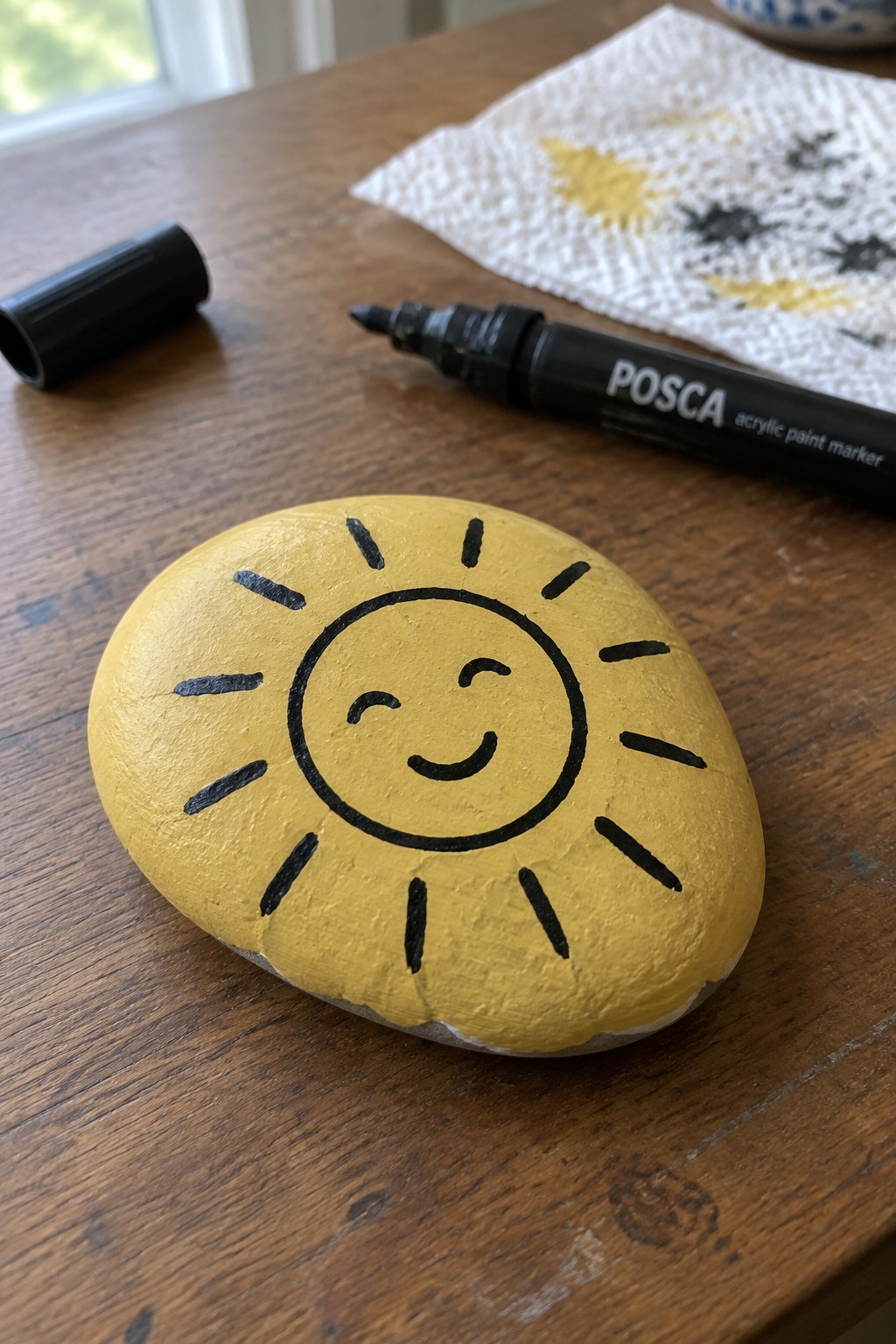 Yellow painted rock with smiling sun face beside black Posca marker on wood.