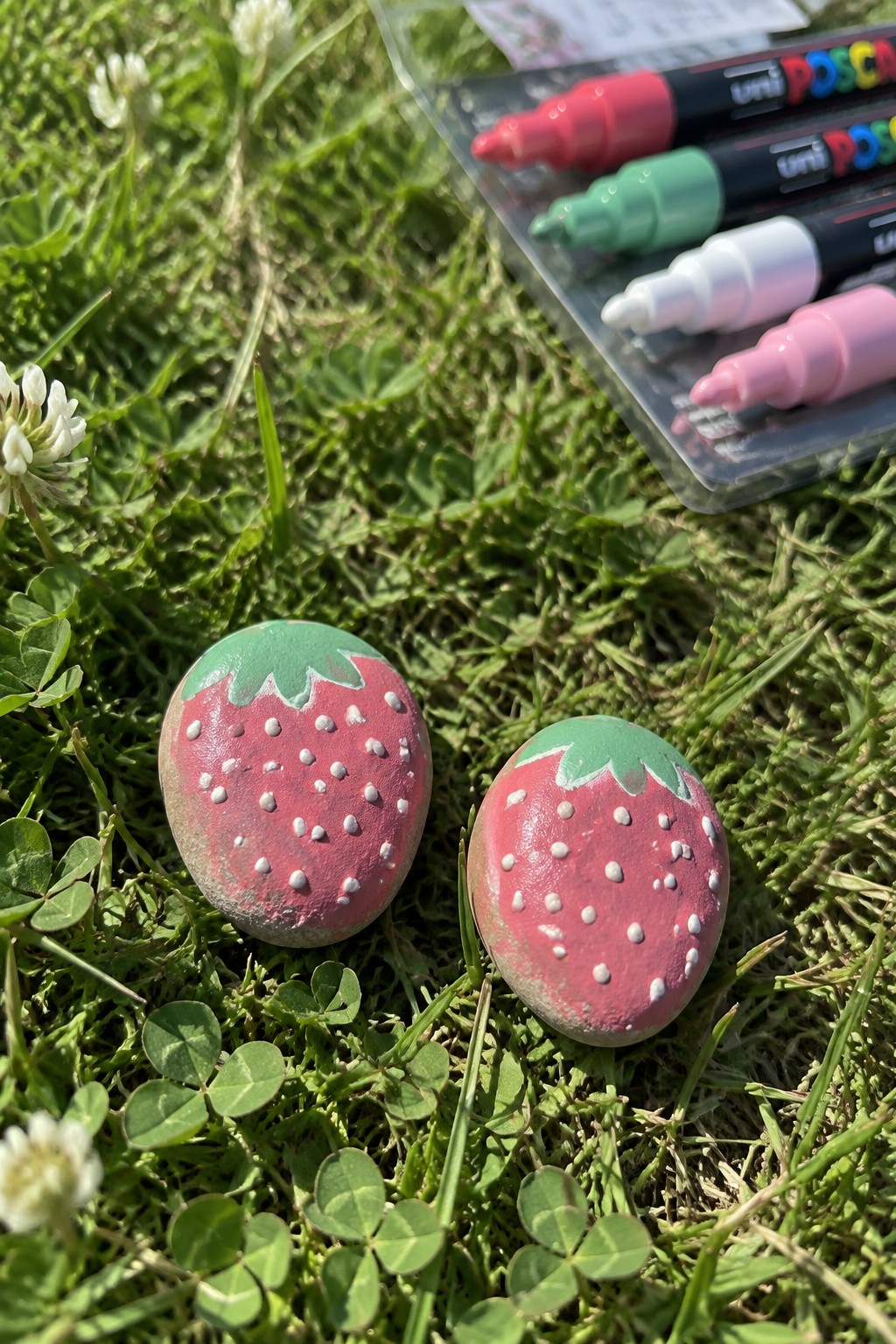 Two strawberry-painted rocks on grass beside colorful Posca markers