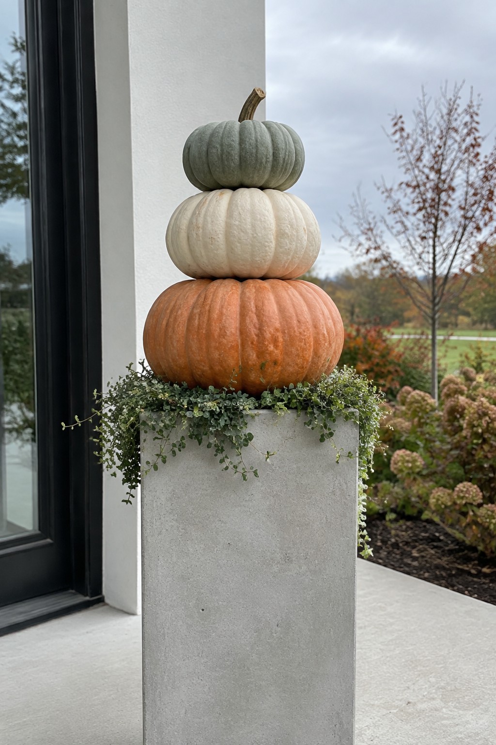 Three stacked pumpkins in green, white, and orange sit on a concrete pillar beside a black door, with greenery draped around the base.