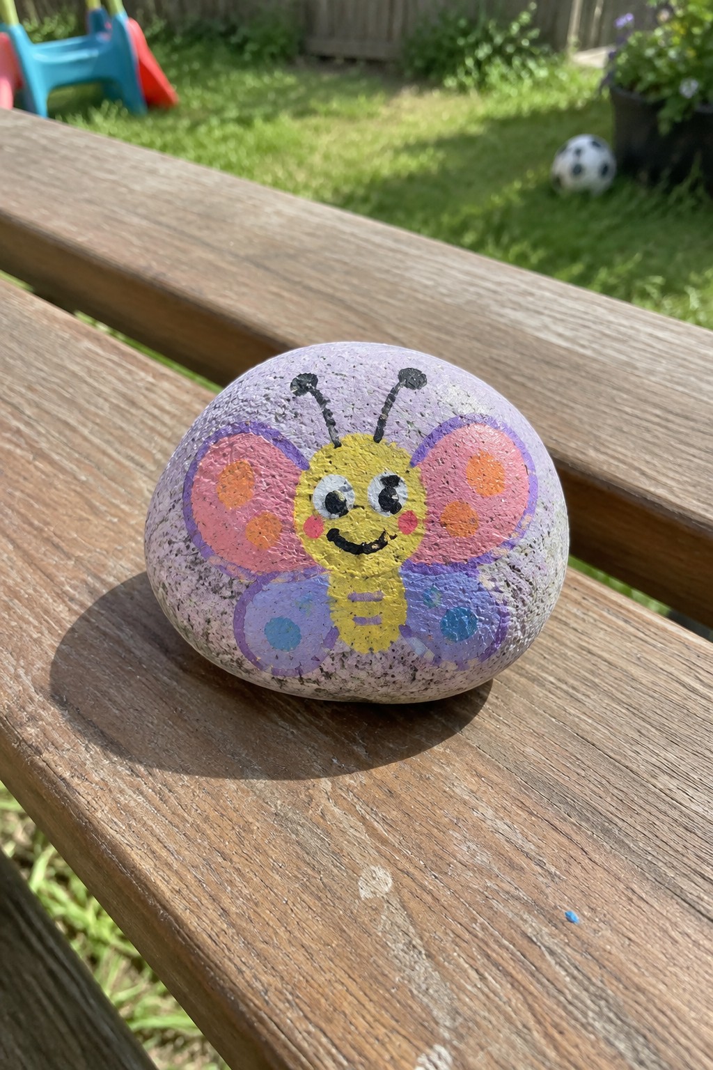 Hand-painted butterfly rock with yellow face and pink-purple wings on bench