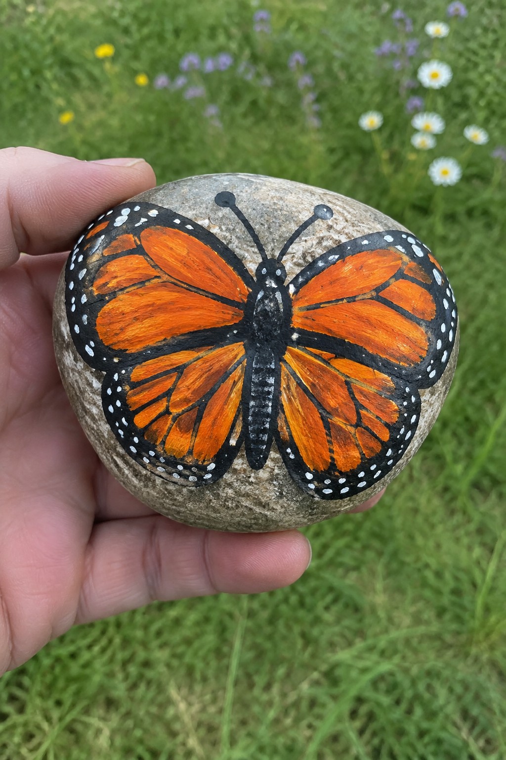 Hand holding stone painted with orange monarch butterfly in grassy field.