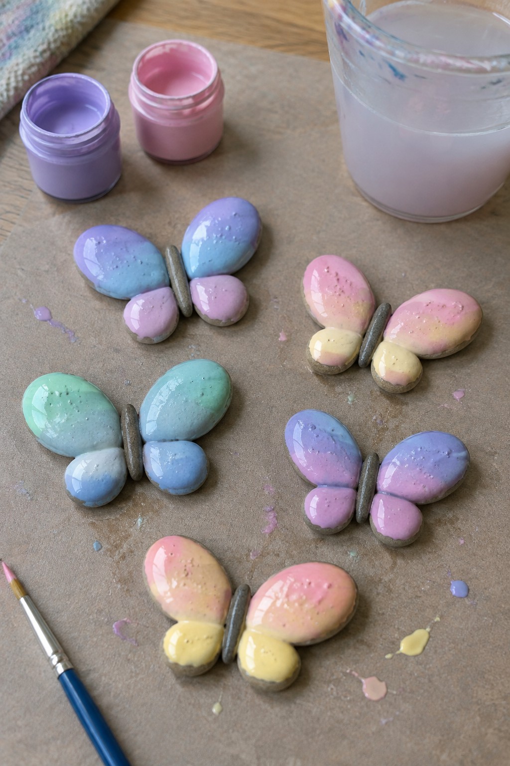 Five pastel gradient-painted stone butterflies with paint jars and brush on paper