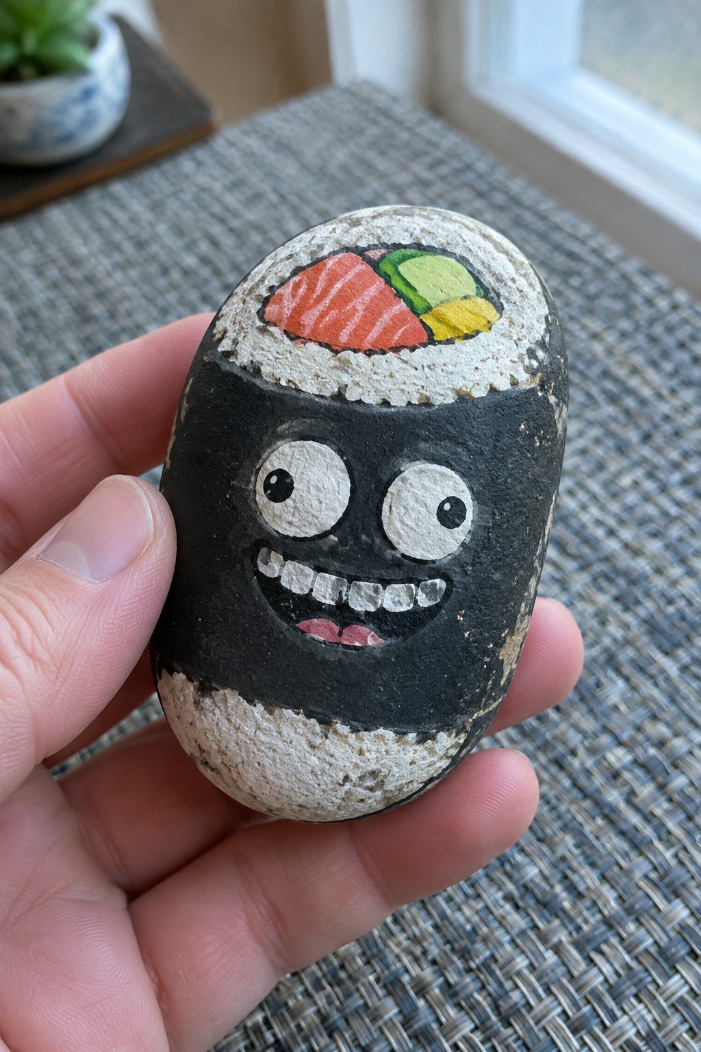 Hand holding rock painted as sushi roll with cartoon face and toppings
